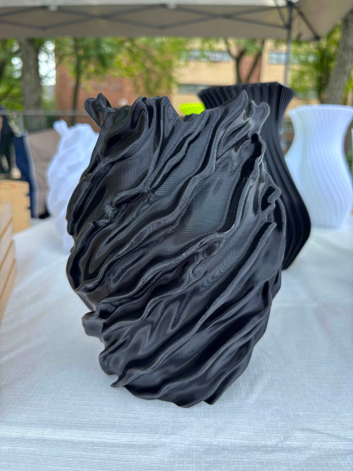 A black sculptural vase with a highly textured, wavy, pleated surface, displayed on a white tablecloth at an outdoor market or gallery event.