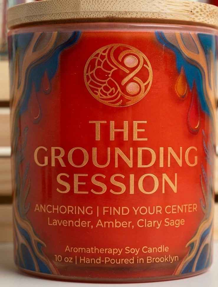 Close-up of 'The Grounding Session' aromatherapy soy candle with red marbled design, gold logo, and label details in warm lighting