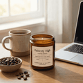 An amber glass jar candle labeled 'Priority High' sits on a wooden desk beside a mug of coffee, a bowl of coffee beans, and an open laptop in a softly lit room.