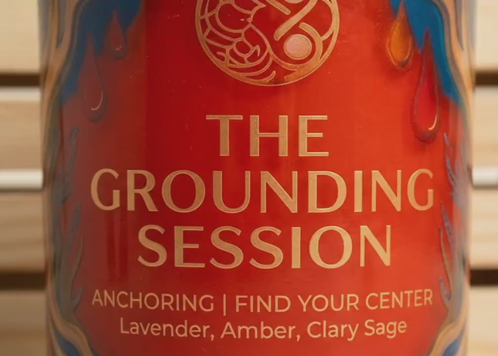 Red aromatherapy soy candle labeled 'The Grounding Session' with gold text, swirling blue and orange accents, and notes of lavender, amber, and clary sage.