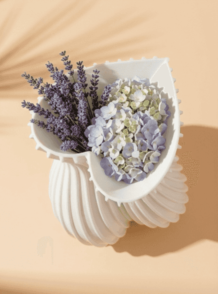 White  sculptural vase with ribbed swirl design, partially open to reveal lavender and hydrangea flowers inside.